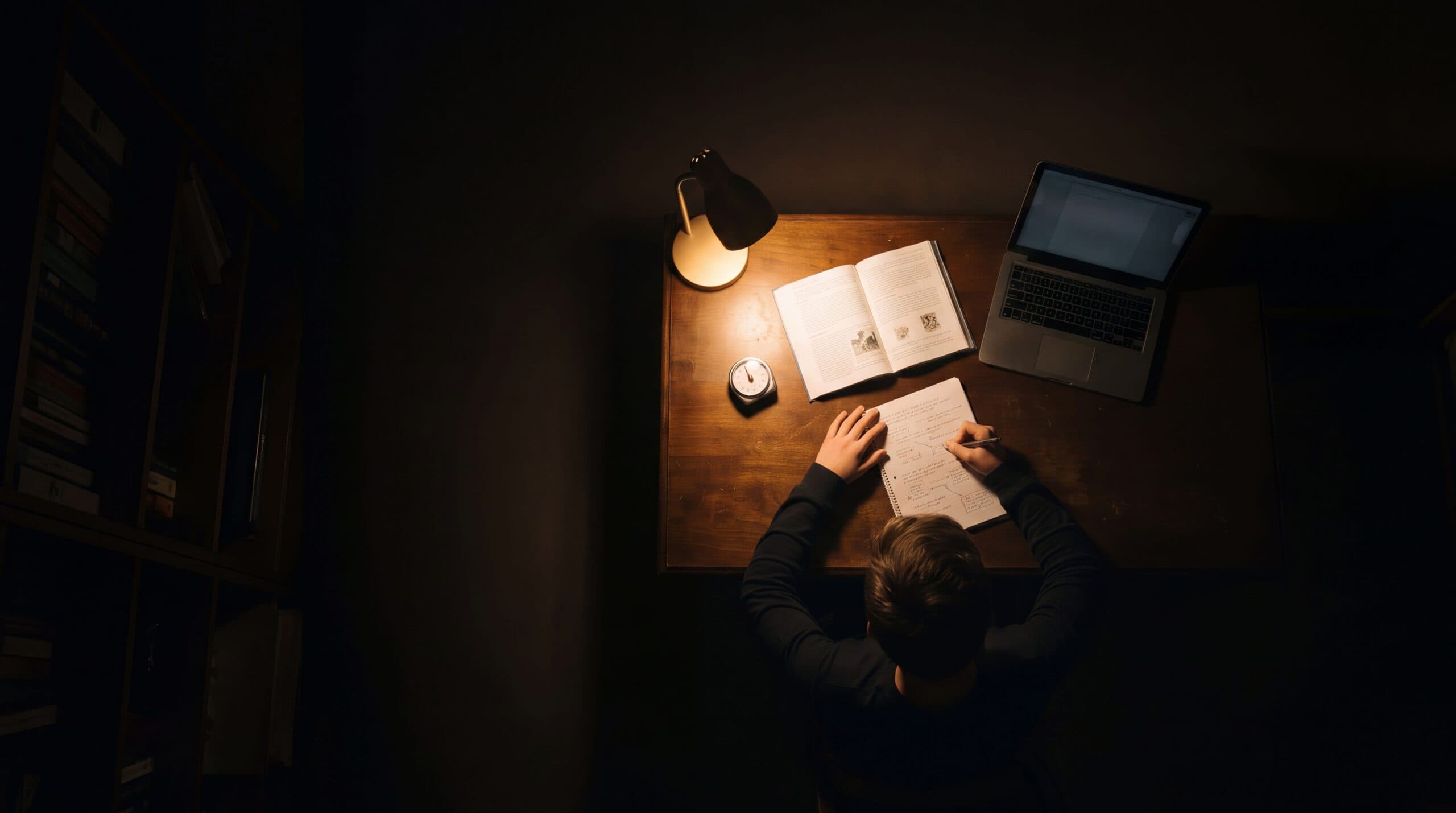Seen from above, a student writes in a notebook under a desk lamp in a dark room.